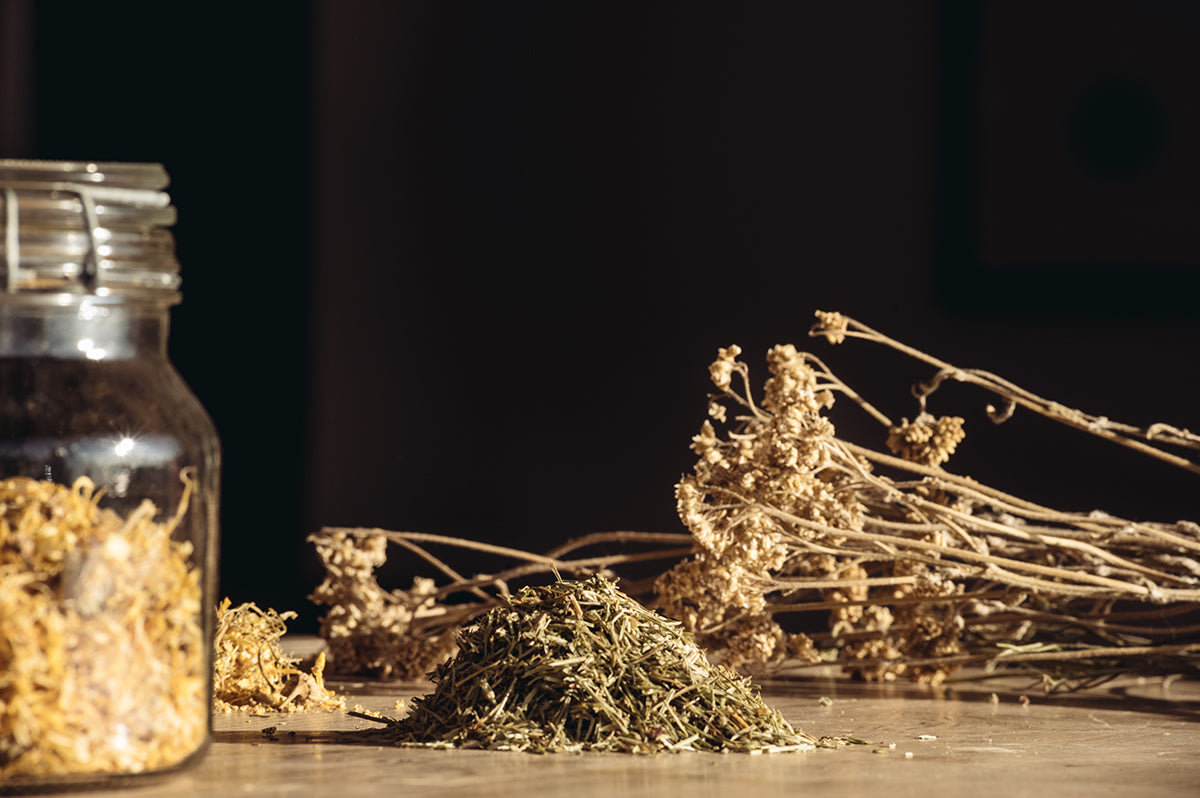 A collection of dried loose-leaf herbs and botanicals spread on a table next to a clear glass jar with a latch lid.